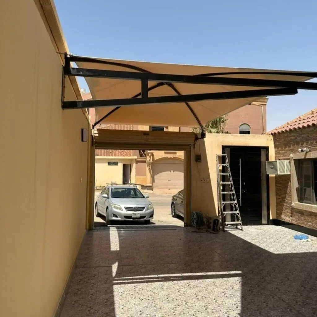 حداد مظلات وسواتر بالرياض 6 ظلة بلون بيج مثبتة على هيكل معدني أسود تغطي مدخل سيارة في منزل، مع سلم بجانب باب أسود وأرضية ببلاط منقوش.
