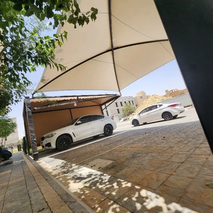 حداد مظلات وسواتر بالرياض 5 مظلة سيارات بيضاء مثبتة على أعمدة معدنية، تغطي سيارة في موقف حضري بجانب الرصيف، مع وجود أشجار ومباني وخلفية إنشائية.