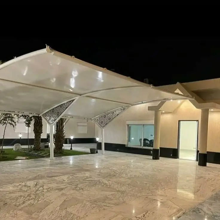 حداد مظلات وسواتر بالرياض 4 مظلة خارجية بيضاء بتصميم هندسي أنيق في منطقة مفتوحة أمام مبنى ليلي، مع أرضية من الرخام ونباتات منسقة وإضاءة خارجية مميزة.