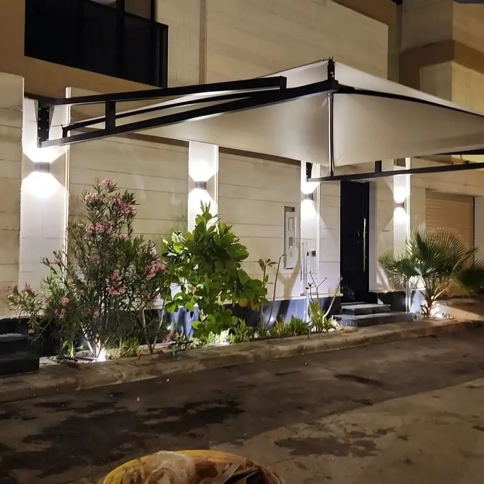 حداد مظلات وسواتر بالرياض 1 واجهة مبنى حديث في الليل مع مظلة خارجية قابلة للسحب بلون أبيض، مدعومة بهيكل معدني أسود، وإضاءة جدارية تبرز جمال التصميم، بالإضافة إلى حديقة منسقة تحتوي على نباتات وزهور.