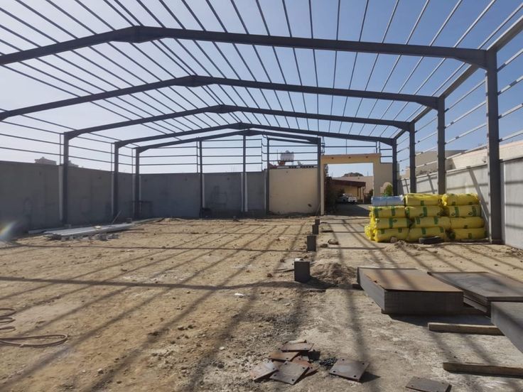 صورة واضحة لهيكل هنجر فولاذي عريض وكبير قيد الإنشاء في منطقة صناعية بالرياض. يظهر الإطار المعدني المتكامل للسقف والأعمدة الرأسية، مع وجود ألواح حديدية سميكة ومواد بناء (أكياس صفراء) مُكدسة على أرضية الموقع الترابية