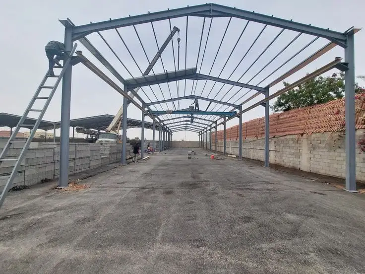 صورة تظهر مرحلة تركيب الهيكل الفولاذي لسقف هنجر في الرياض، حيث يتم تثبيت الجوائز المعدنية بواسطة رافعة، ويُشاهد عمال بناء يعملون على الهيكل، مع وجود بلوك أسمنتي مكدس في الخلفية.