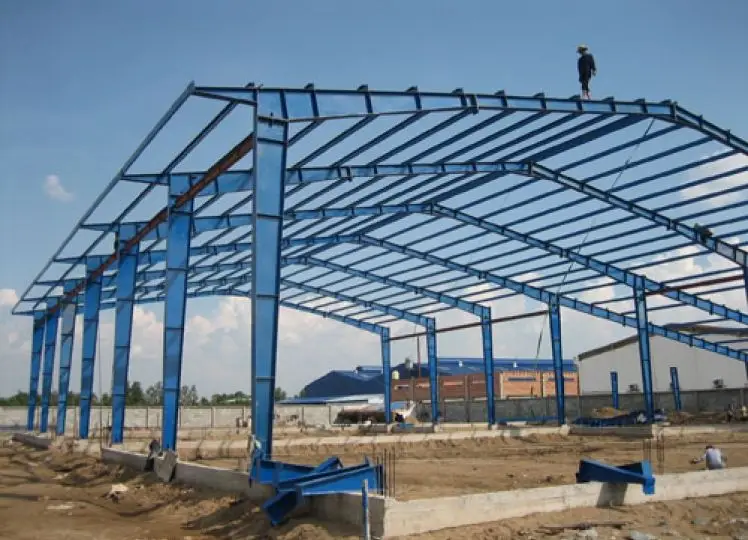 صورة لهيكل هنجر فولاذي مكتمل الإطار ومطلي باللون الأزرق الساطع، يرتكز على قاعدة خرسانية مرتفعة. يظهر عامل بناء يقف على قمة السقف، مع مبانٍ صناعية أخرى في الخلفية.